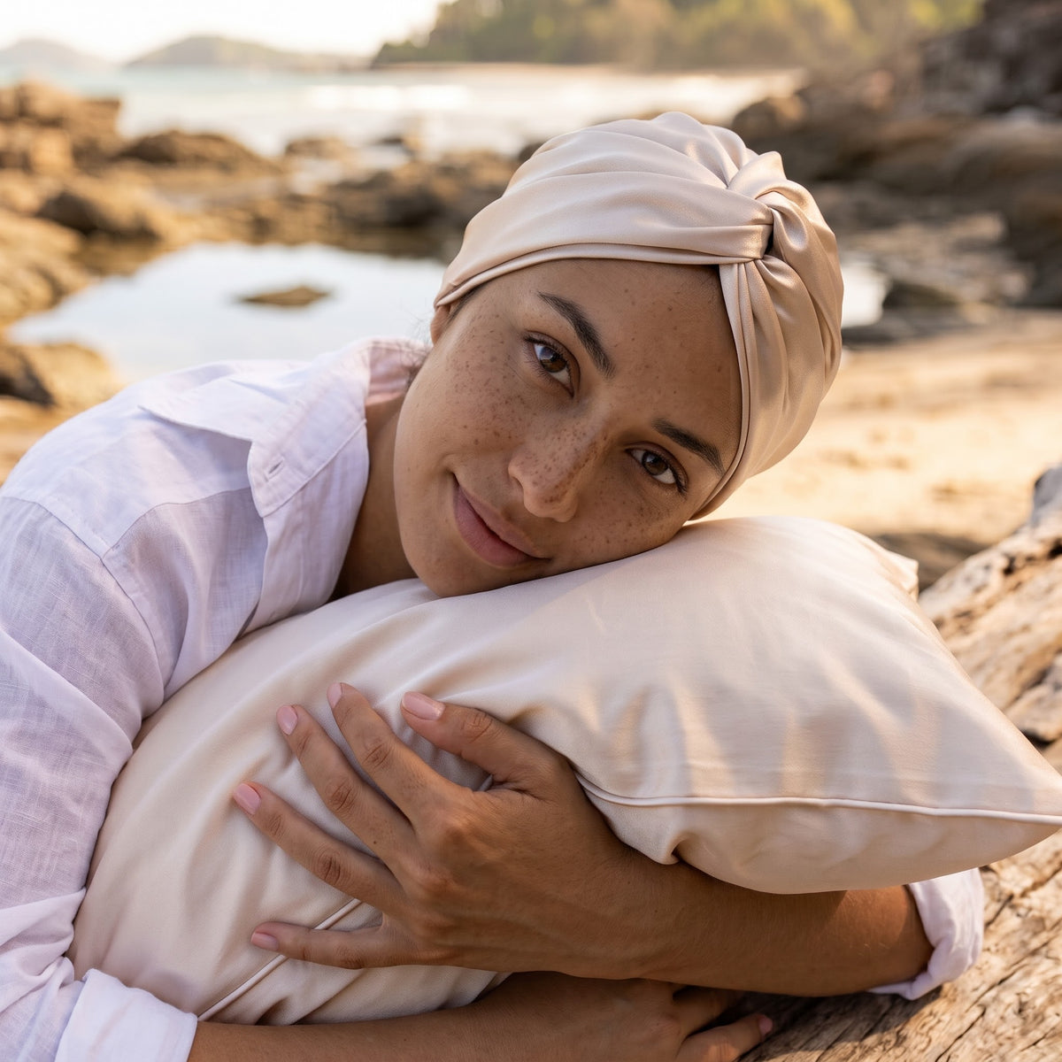 A woman with a beige headwrap and white shirt rests her head on a matching beige pillow on a rocky beach.